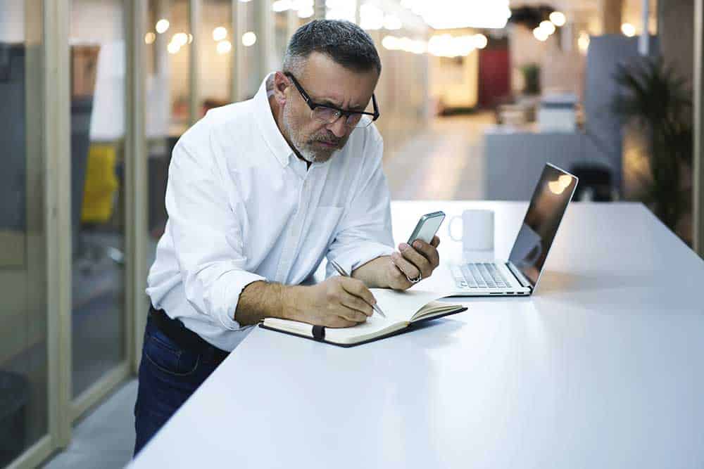 Mann im Büro schreibt etwas in sein Notizbuch und hält mit der anderen Hand sein Smartphone.